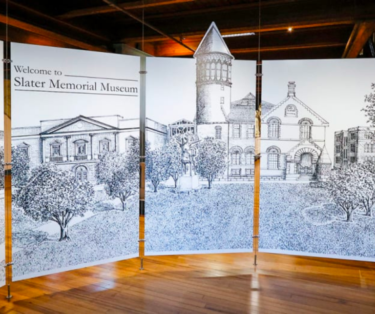 A panel of canvases stand freely in a large room. The canvases have black and white line drawings of the Slater museum.