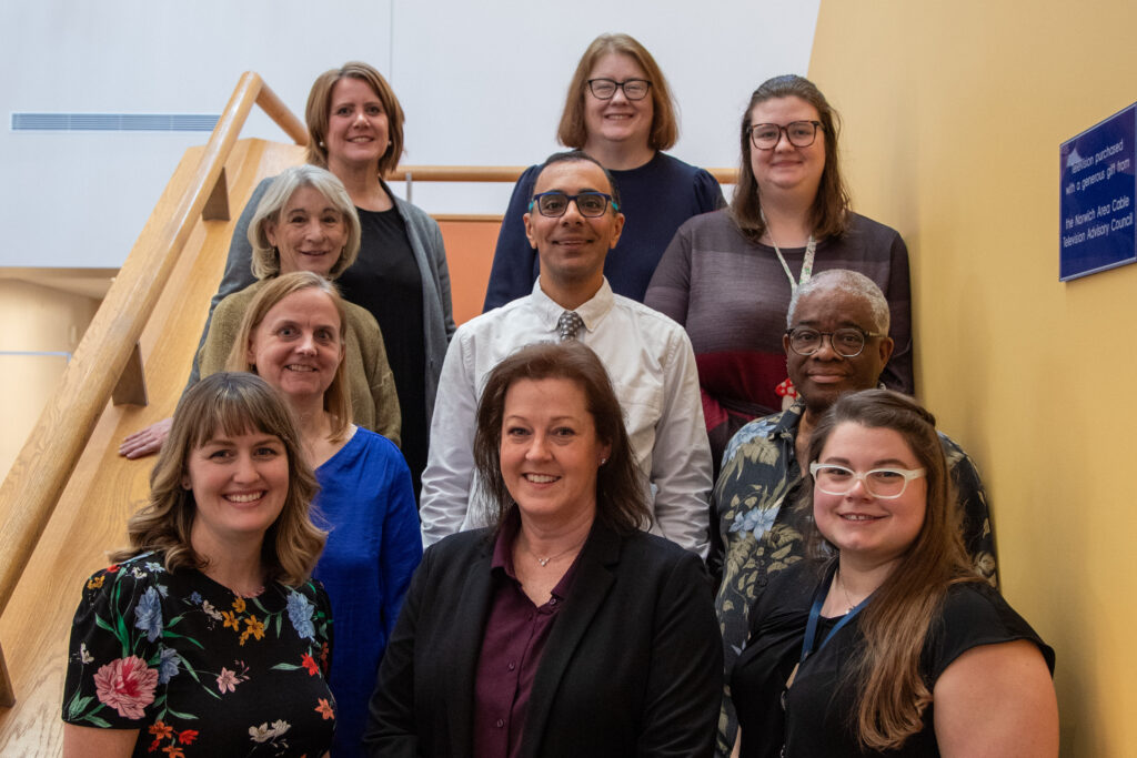 The staff stands on the stairway in the atrium. Three are on each step, making it so that all faces can be seen. They are smiling at the camera, inviting you in to visit Otis Library!