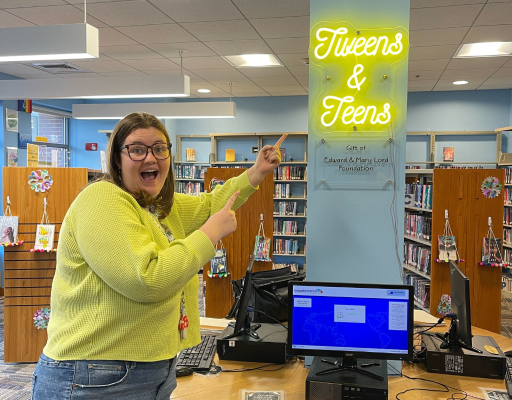 Kait pointing at the sign welcoming teens and tweens to Otis Library