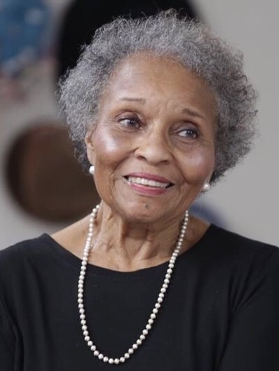 Lottie B. Scott smiles softly at someone off screen. She is wearing a black top with pearl earrings and a necklace. She looks elegant and dignified.