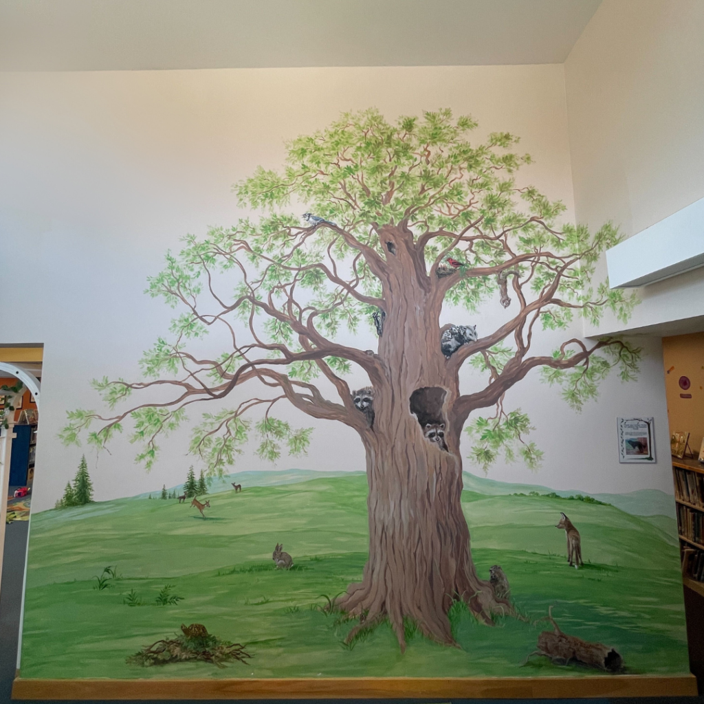 A large tree stretches from floor to ceiling. Its branches are home to little native woodland creatures. Green fields reach about half way up the wall.