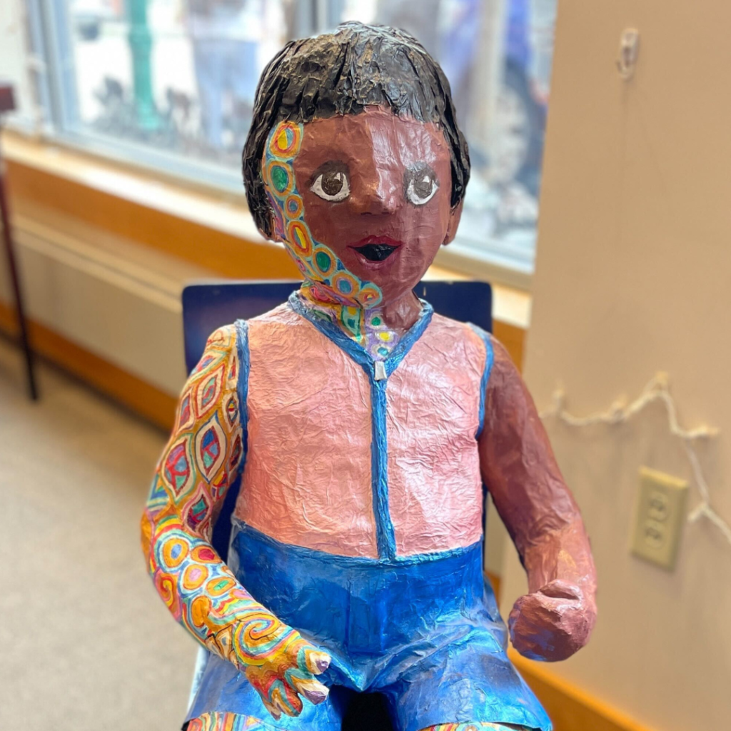 The Papier-Mâché child wears a peach and blue tank top. His dark brown skin is accented with small, confetti-like pieces of color. The colors represent mental illness, specifically schizophrenia.