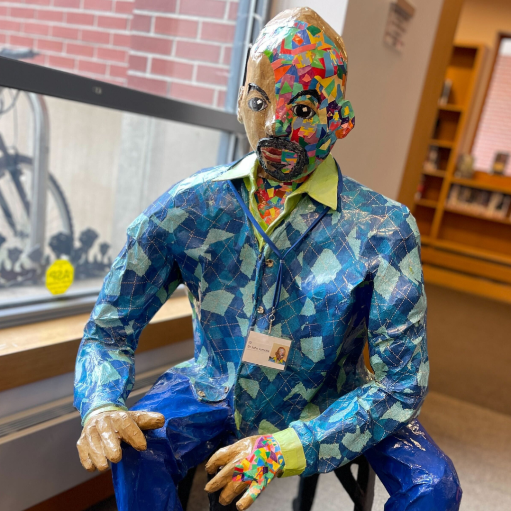 The Papier-Mâché Doctor wears a blue shirt of many shades in a seemingly random pattern. His light brown skin is accented with small, confetti-like pieces of color. The colors represent mental illness, specifically schizophrenia.
