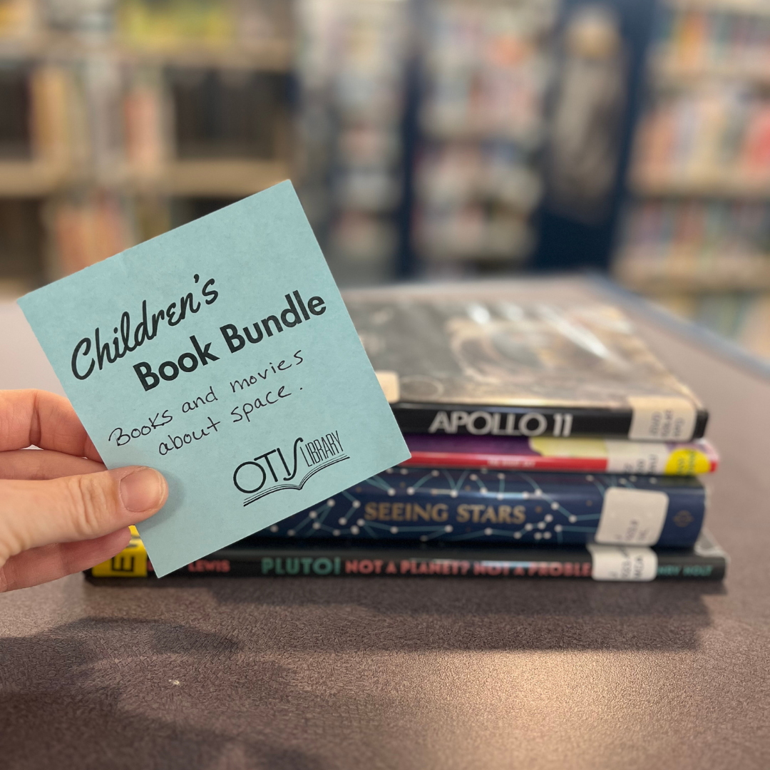 A hand holds up a blue card in front of a stack of books. The card reads "Children's Book Bundles." In handwriting underneath is a list of topics that match the books