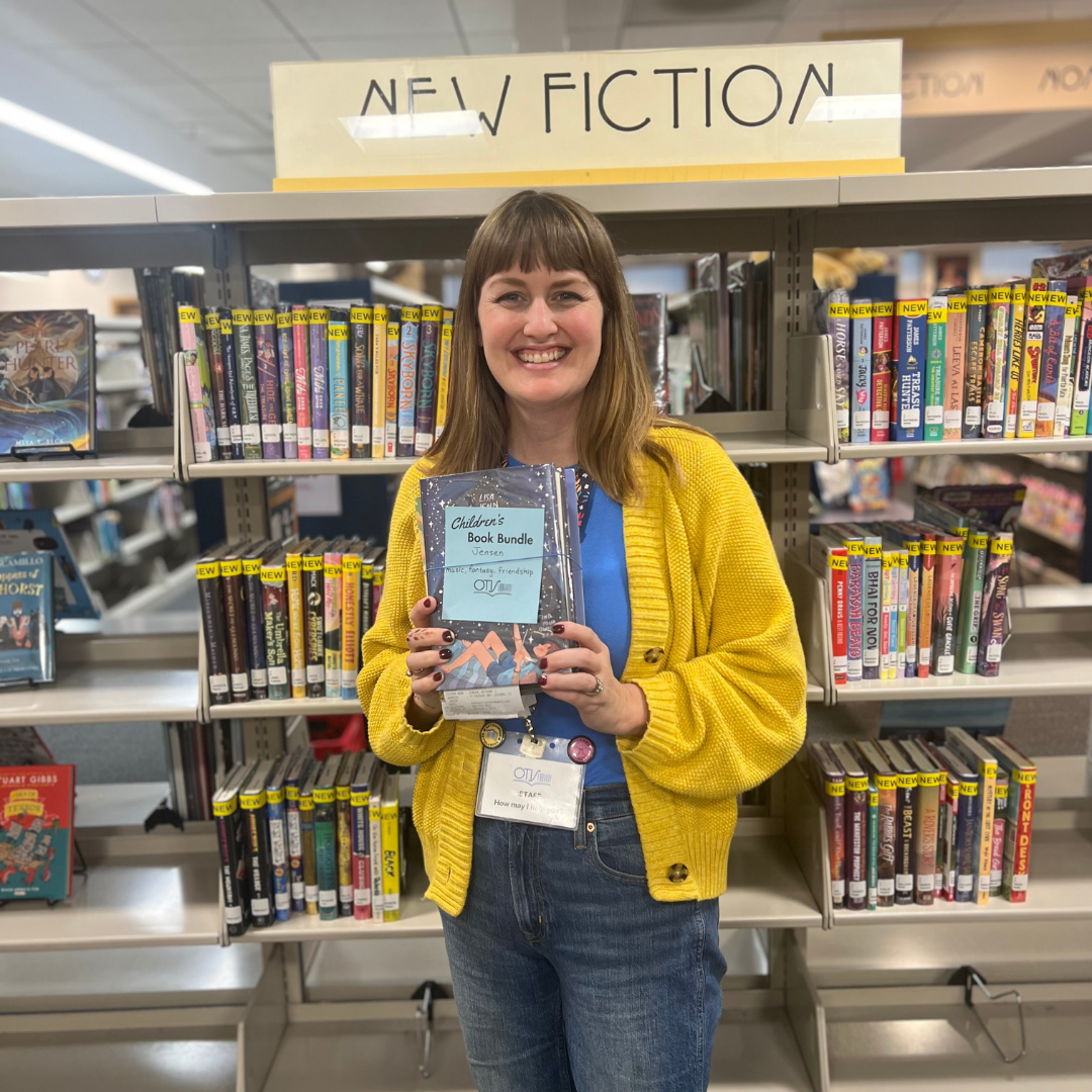 Bethany smiles at the camera as she holds up a stack of books. A blue card is taped to the cover, identifying it as a book bundle. She is very excited about the new service!