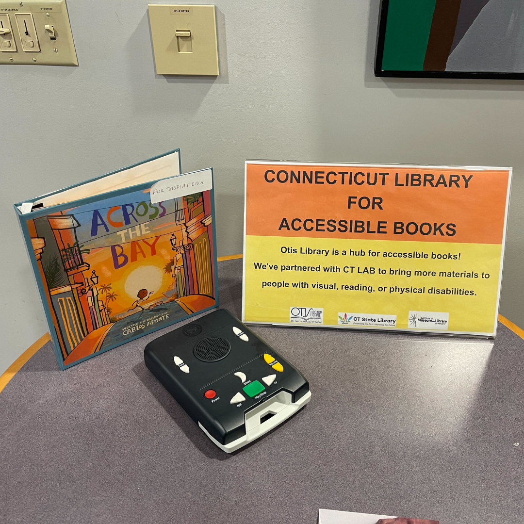 A table displays multiple items from CTLAB, including a children's book in braille, a "Talking Book" player, and a sign explaining the program. The display is open to the public and inviting people to explore the items.